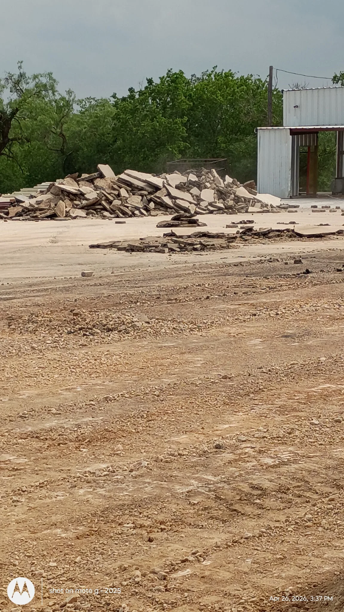 Demolished concrete and masonry debris piled on cleared dirt lot with metal storage building